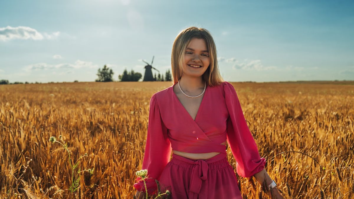 A smiling woman in a pink outfit standing in a sunlit wheat field with a windmill in the background.